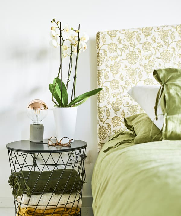 Letto con biancheria verde e testiera in tessuto fantasia cachemire verde, un paio di occhiali e un'orchidea su un comodino in metallo intrecciato.
