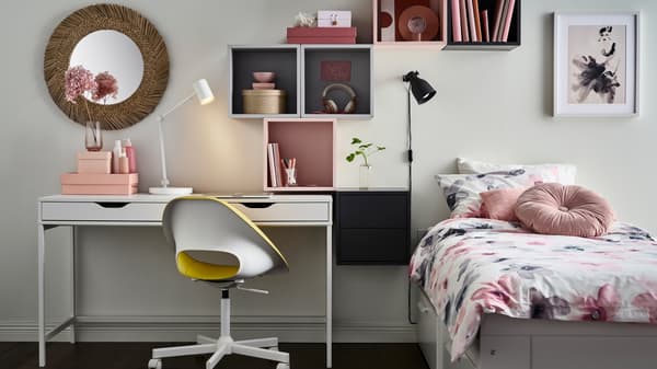 Letto divano BRIMNES bianco con biancheria LAPPNYCKLAR fantasia, accanto a una scrivania ALEX bianca con mobili grigi e rosa sopra - IKEA.