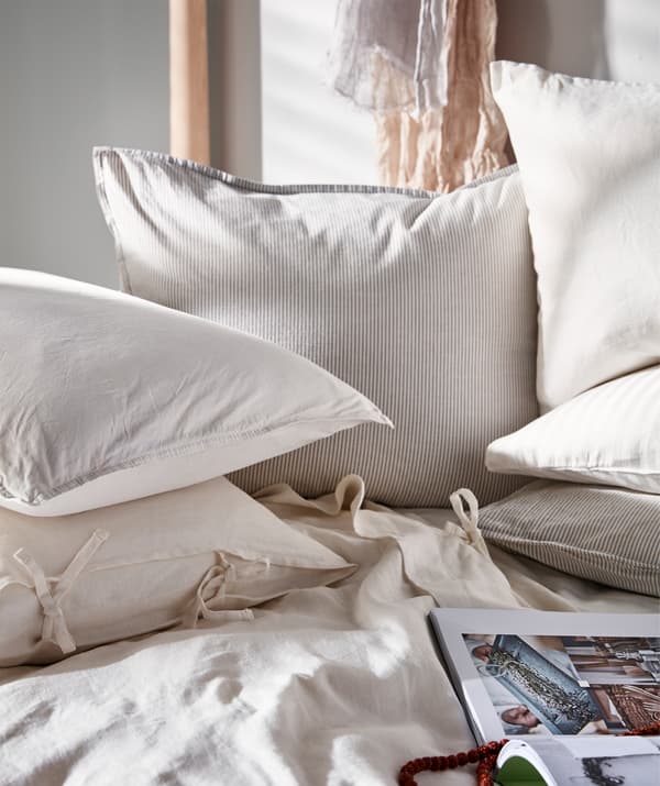 Letto illuminato dal sole con cuscini in tessuto chiaro, un libro aperto appoggiato al piumino - IKEA