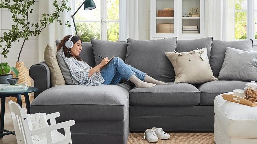 Lezy na szarej narożnej kanapie. Ma słuchawki na głowie, na kolanach notes, w którym notuje.