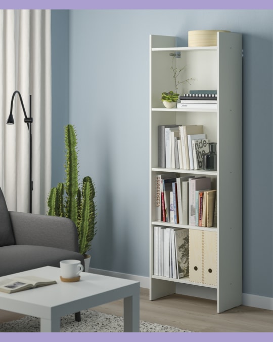 Librero blanco de cinco niveles en un living, con libros, archivadores y una planta decorativa en la repisa superior.