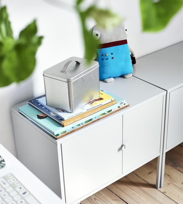 Libri appoggiati sopra un mobile grigio insieme a un pupazzo e un contenitore di metallo. Sulla sinistra si intravede una tastier a un tappetino di un mouse.