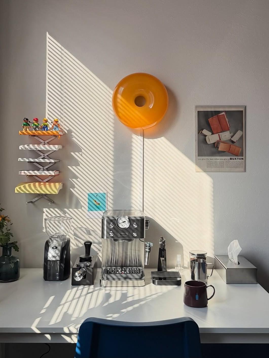 Lichte koffiecorner met een espressomachine op een wit bureau, koffiemolen en kopjes, een felgele wandlamp, decoratieve plankjes en zonlicht dat strepen werpt op de muur.
