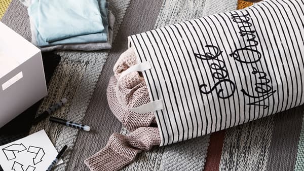 Lien vers « Étape 3 - Détachez-vous de vos choses » - photo d’un fourre-tout en toile rempli de vêtements avec les mots « Seek new owner » (Cherche nouveau propriétaire) brodés dessus, posé sur un tapis près de boites, de stylos et d’une étiquette de recyclage.