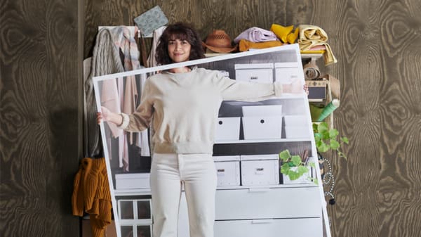 Lien vers « IKEA Harmoni - Désencombrez votre vie » - photo d’une femme habillée en blanc, debout devant des étagères encombrées et tenant en main une grande photo de ces mêmes étagères bien rangées.