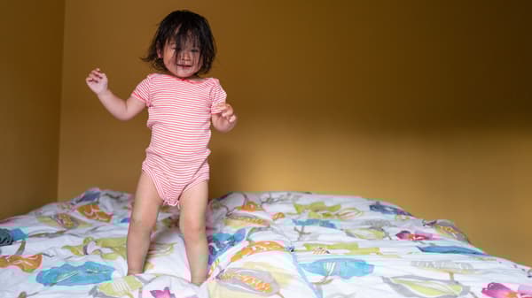Lien vers « La vie à la maison en des temps incertains - Partie 1 : changements » - image d'une petite fille aux cheveux foncés couvrant son visage, sautant joyeusement sur un lit.