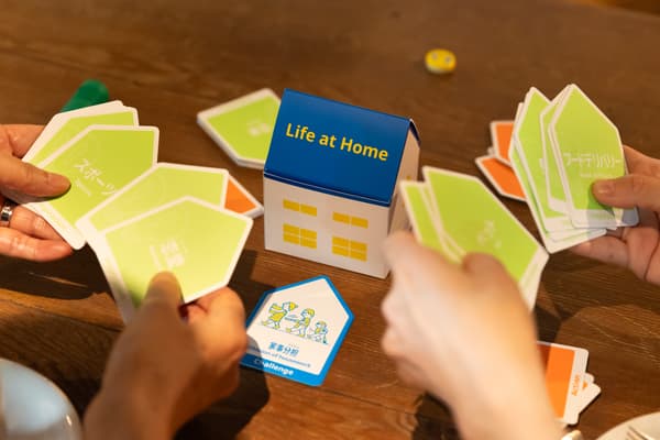 『Life at Home』と書かれた家型のボックスを囲む人々が、緑のカードを手に持っている様子。