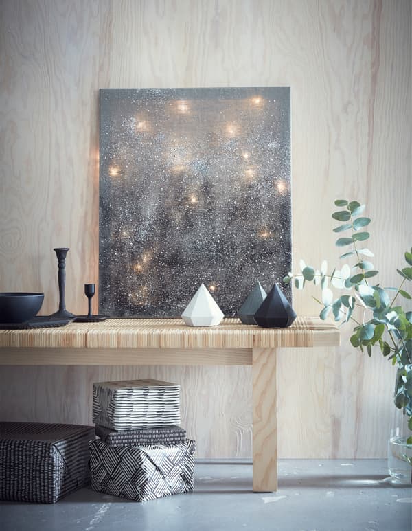 Light chains arranged behind a grey canvas, leaning on a light wood bench, with ornaments and gifts.