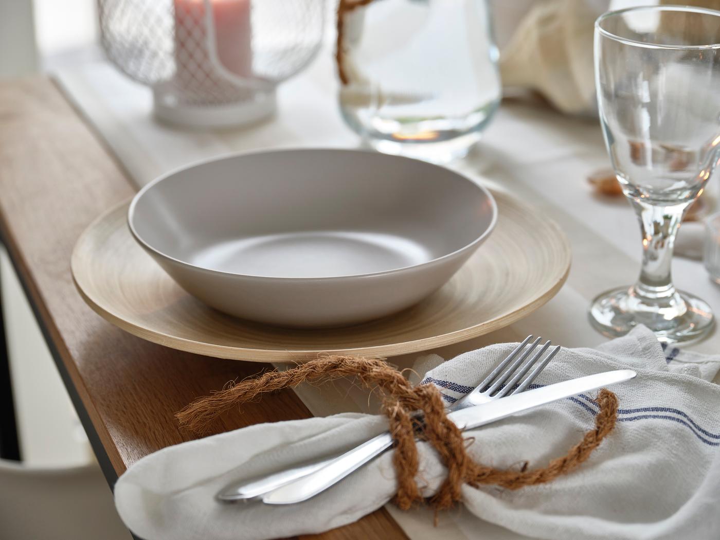 Light table setting with bowl on wooden charger plate, glass, tealight and napkin with cutlery.