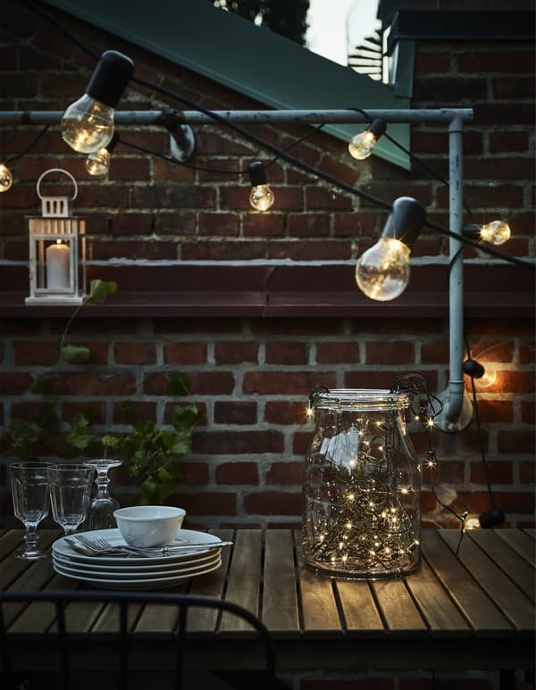 Lighting chains, a lantern, some glasses and plates and cutlery on a table on a terrace.