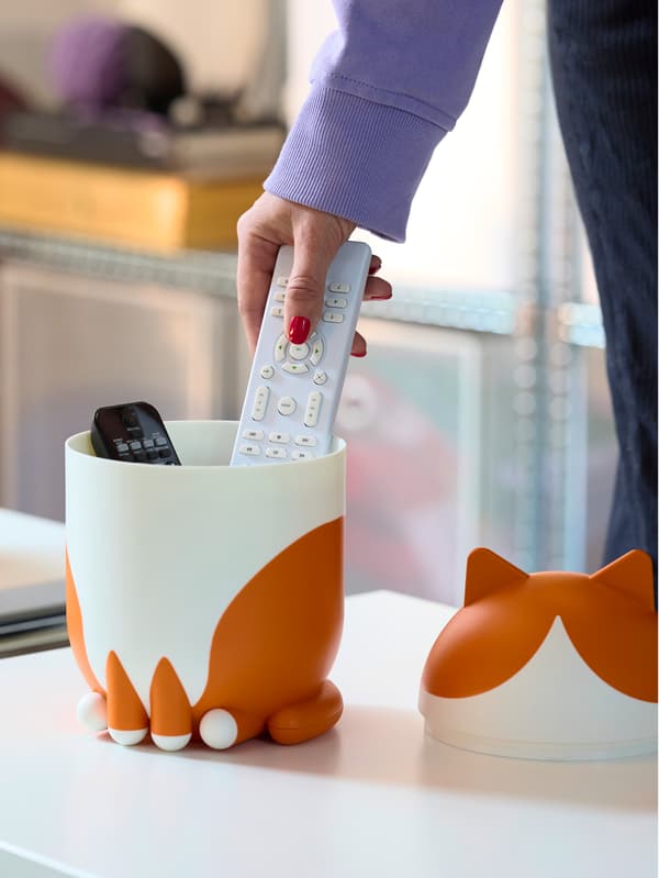 Lila pulóveres kéz egy fehér távirányítót tesz a fehér/narancssárga cicaformájú GREJSIMOJS tárolóba.