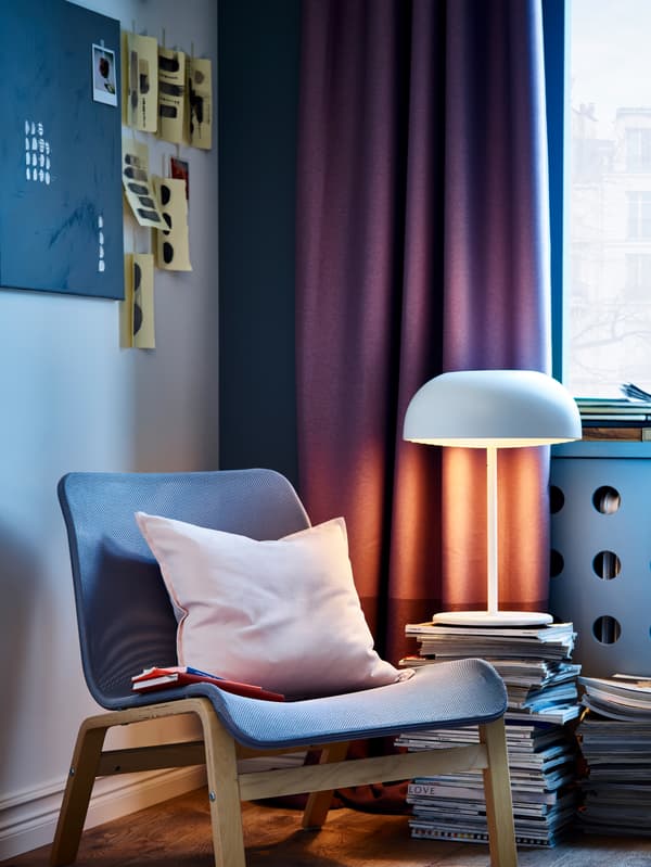 Lilac curtains, a white table lamp, a light pink cushion cover and an easy chair in grey and birch veneer.