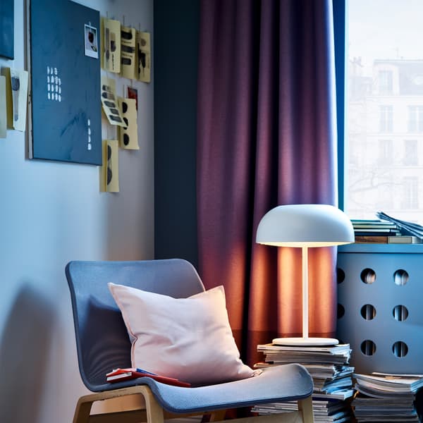 Lilac curtains, a white table lamp, a light pink cushion cover and an easy chair in grey and birch veneer.