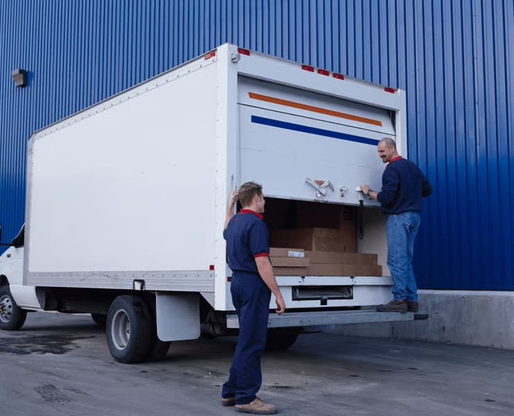 L’image montre deux livreurs en train de charger ou décharger un camion blanc garé devant un bâtiment bleu, typique d’un entrepôt IKEA. On aperçoit à l’intérieur du véhicule plusieurs colis plats et cartons, caractéristiques des emballages de meubles. La scène illustre la logistique et l’organisation derrière la livraison, dans un cadre professionnel et structuré.