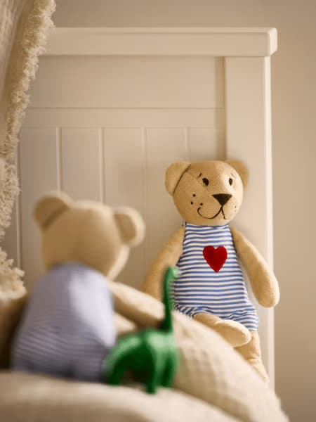 L’image montre deux oursons en peluche dans une chambre d’enfant, l’un en arrière-plan assis contre un lit blanc, portant une salopette rayée avec un cœur rouge, l’autre flou au premier plan avec un petit dinosaure vert. L’ambiance est douce, chaleureuse et rassurante, évoquant la tendresse de l’enfance et un moment de jeu paisible.