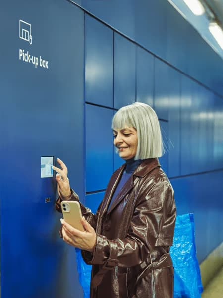 L’image montre une femme souriante en train de récupérer un colis dans une « Pick-up box » bleue, typique d’un service de retrait automatique. Elle tient son téléphone dans une main et interagit avec l’écran du casier avec l’autre. En arrière-plan, un grand sac bleu IKEA dépasse de son dos. La scène illustre la simplicité et la praticité d’un service de retrait en libre-service, rapide et autonome.