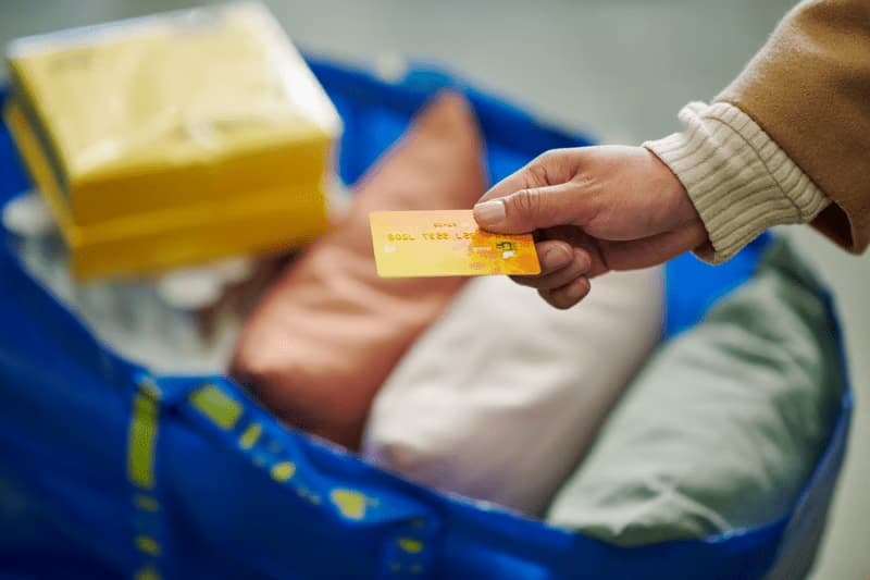 L’image montre une main tendant une carte bancaire orange, prête à effectuer un paiement. En arrière-plan, un grand sac bleu IKEA rempli d’articles textiles (coussins colorés) et de boîtes jaunes suggère une séance de shopping. La scène illustre un moment d’achat simple, pratique et quotidien, dans un univers IKEA reconnaissable.