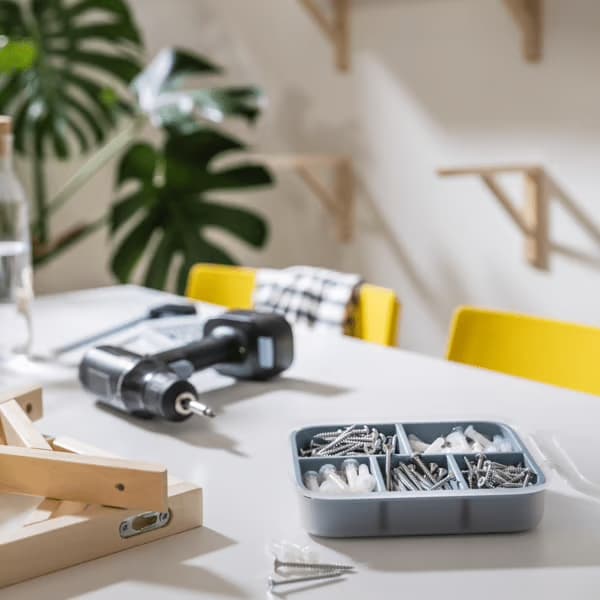 L’image montre une scène de bricolage en cours sur une table blanche. On y voit une perceuse, une boîte compartimentée remplie de vis et chevilles, quelques pièces de bois et des outils. À l’arrière-plan, des chaises jaunes et une grande plante verte apportent une touche de couleur et de vie. L’ensemble évoque un moment de montage ou d’aménagement, dans un intérieur lumineux et organisé.
