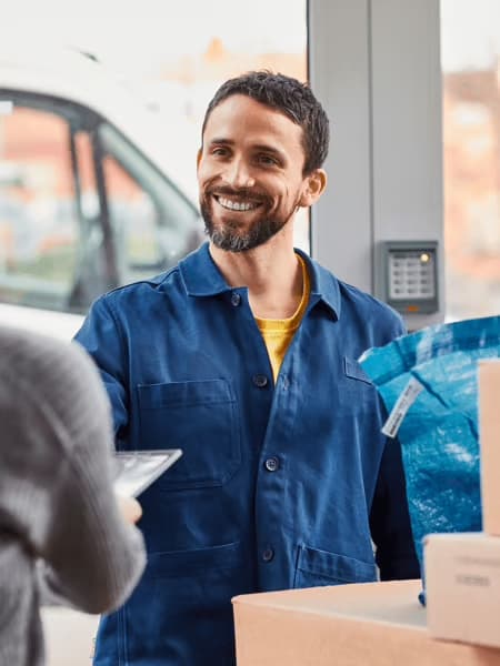 L’image montre une scène de livraison ou de retrait de colis dans une ambiance conviviale. Un employé souriant, vêtu d’une veste bleue, échange avec une cliente tenant une tablette. Autour d’eux, plusieurs cartons et un grand sac bleu IKEA évoquent un service efficace et chaleureux, centré sur la satisfaction client.
