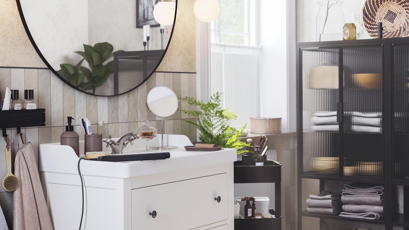 LINDBYN tükör lóg egy fehér, kétfiókos HEMNES/RÄTTVIKEN mosdóállvány felett, egy FRIHULT fali lámpa mellett.