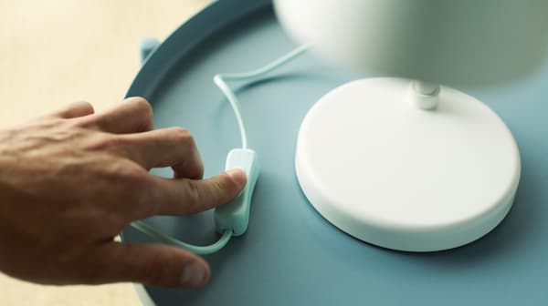 L’index d’une personne sur l’interrupteur d’une lampe de bureau à LED FUBBLA posée sur une table ronde bleu clair.
