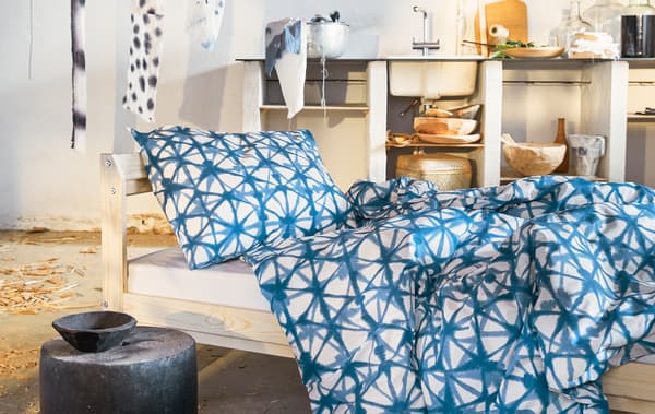 Linge de lit bleu à motif posé sur un lit avec cadre en bois dans une pièce avec des étagères ouvertes et un évier.