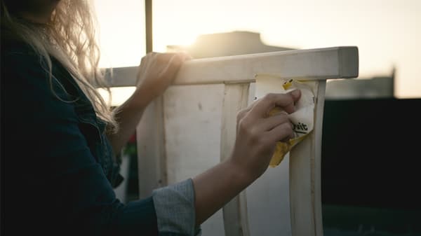 Link to 'Renew it' - image of hands holding sandpaper and sanding a white-painted wooden chair outside with sun glare.