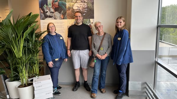 Links im Bild steht ein IKEA-Wagen mit Pflanzen und weiteren Produkten. In der Mitte stehen vier Personen im IKEA-Einrichtungshaus. Zwei der Personen tragen blau-schwarz gestreifte IKEA-Oberteile und die anderen Alltagskleidung.