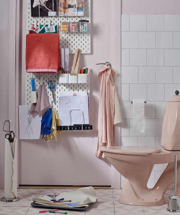 L’interno di un bagno con un pannello portaoggetti appeso alla porta contenente penne, fogli da disegno e altra cancelleria; fogli e penne vicino al water.