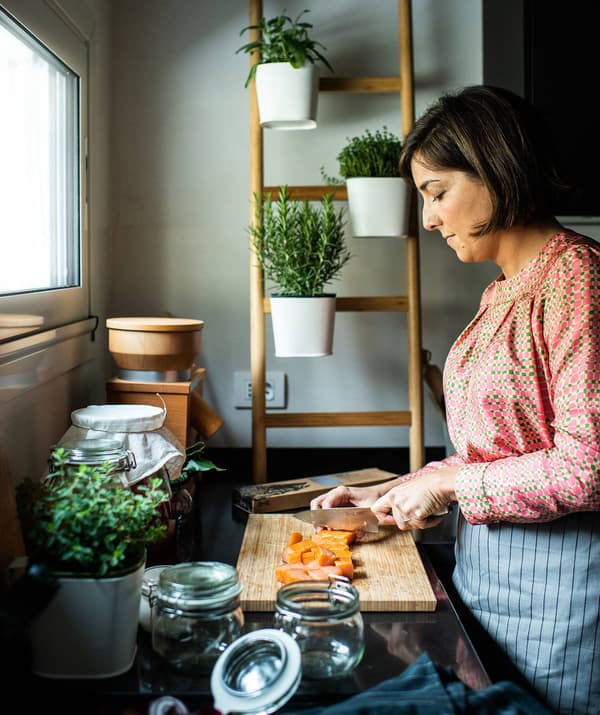 Lisa Casali con un grembiule mentre pulisce e taglia verdure con un coltello in cucina. Sullo sfondo un supporto da parete SATSUMAS con contenitori per piante.