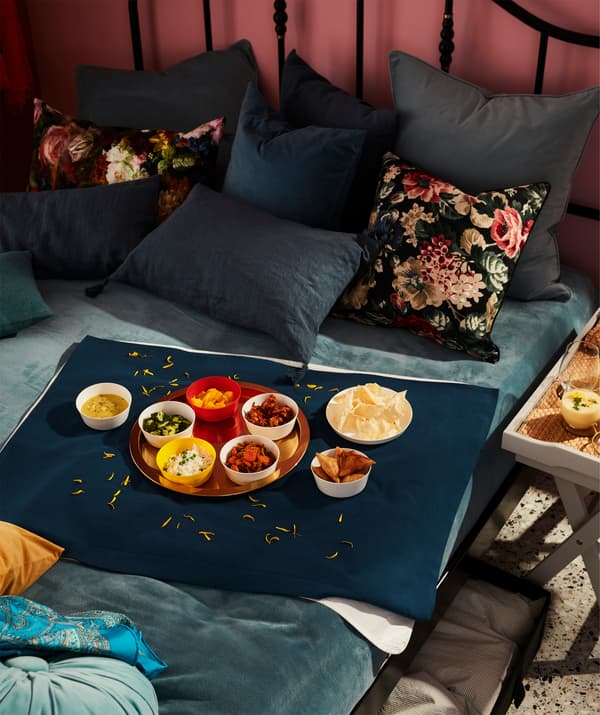 Lit recouvert d'une couverture et beaucoup de coussins sur lequel est servi un repas posé au milieu d'un morceau de tissu qui sert de nappe.