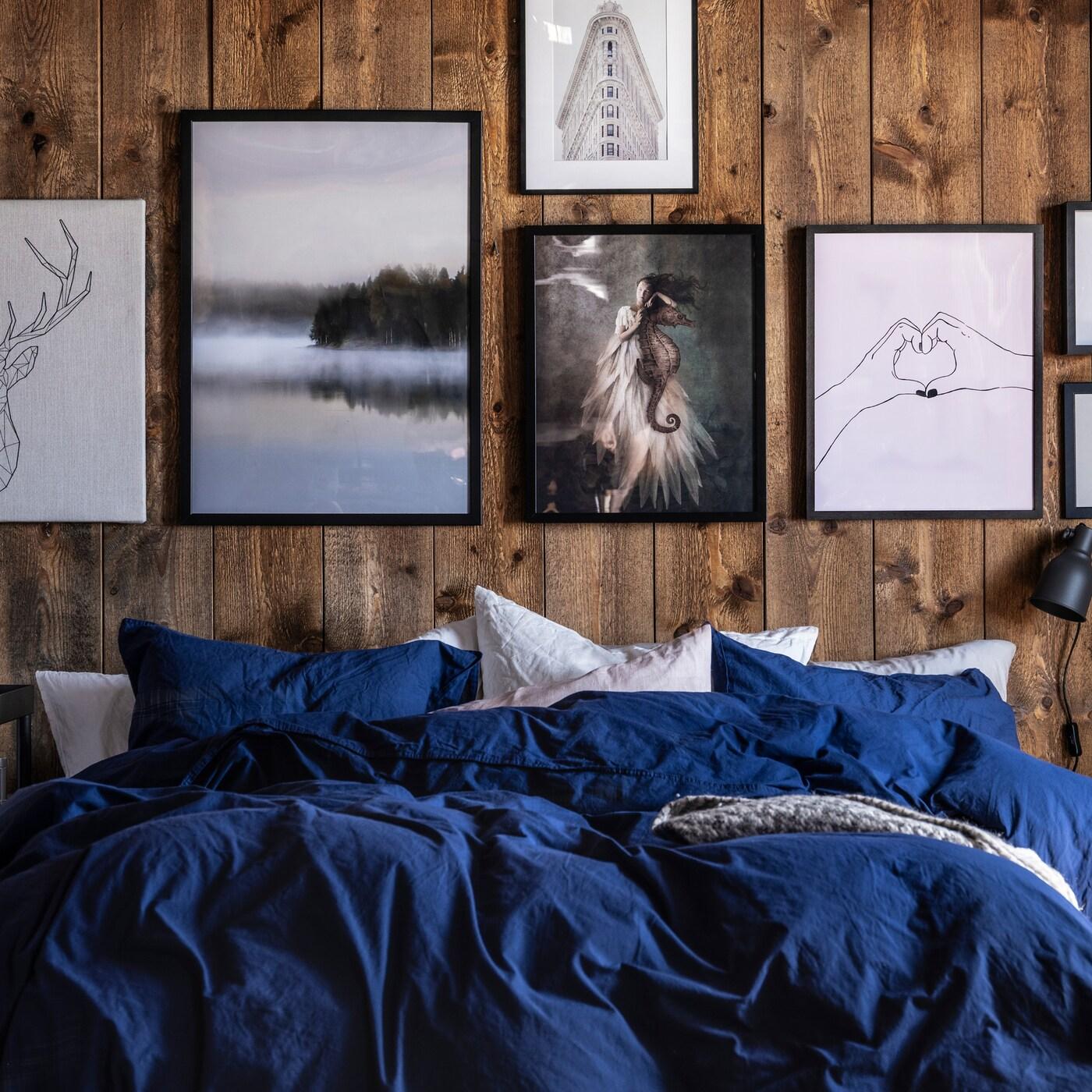 Lit revêtu de linge de lit bleu/blanc devant un mur en bois orné de différentes images dans les tons noir, blanc et gris.