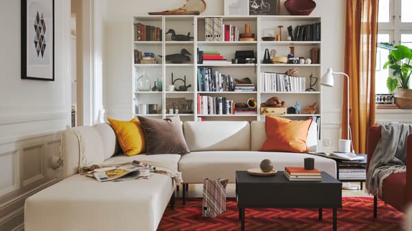 Living con un sofá en L con cojines de diferentes colores sobre el. Detrás del sofá hay un estante con libros y diferentes artículos de decoración.