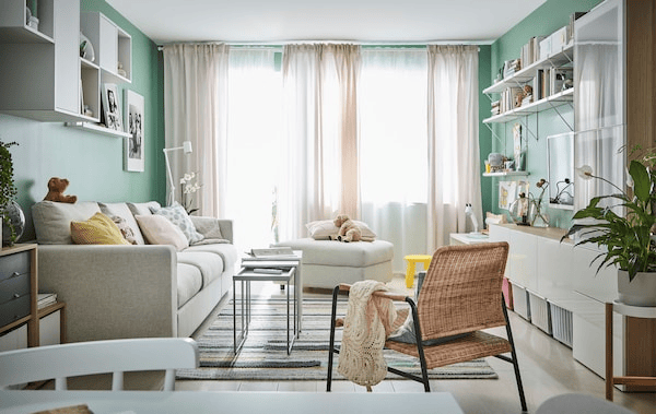 Living room lit up through sheer curtain hanging in front of big windows at far end. Sofa, seating, open storage along walls.