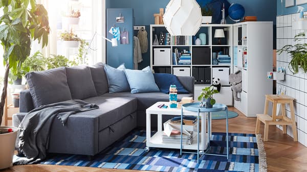 Living room with a dark gray corner sofa-bed, a blue-patterned rug and a freestanding shelving unit with space behind it.