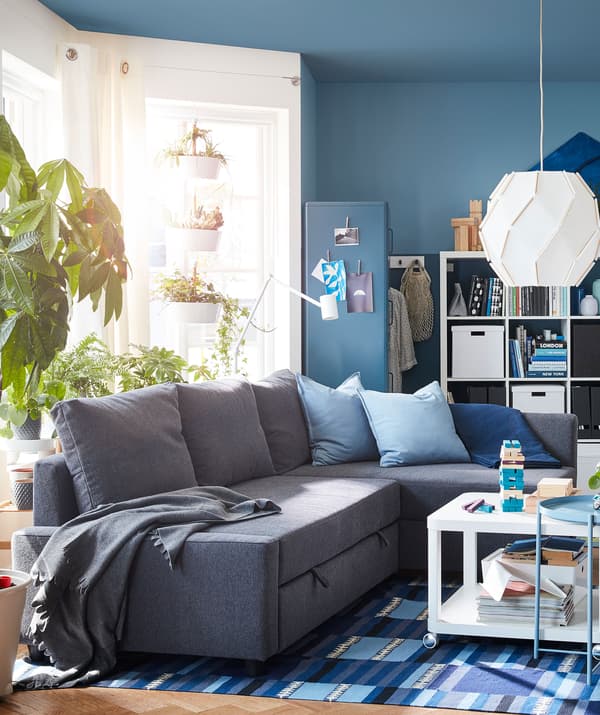Living room with corner sofa in the centre, surrounded by windows, several plants and large bookshelves filled with things.