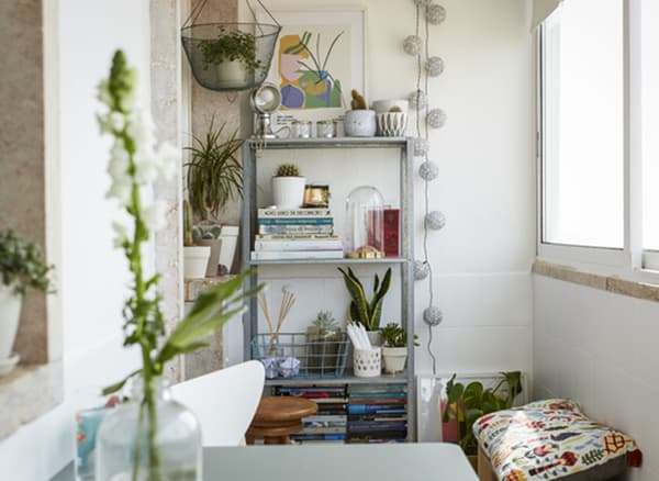 Livros, plantas e peças decorativas numa estante aberta alta, encostada a uma parede branca.