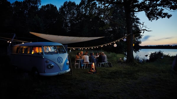Ljudi sede za stolom na otvorenom, pored jezera, pod svetlima i ceradom, s kombijem Volkswagen parkiranim u blizini.