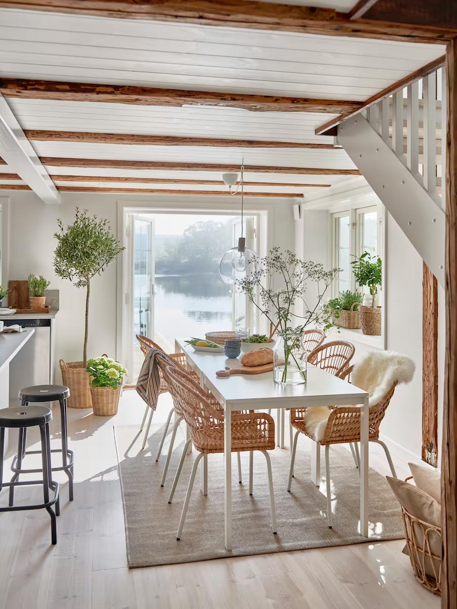 Ljus matsal med vitt bord, rottingstolar och stora fönster med utsikt över vatten. Rummet har synliga takbjälkar, gröna växter och en trappa till höger – en modern och naturnära inredning.
