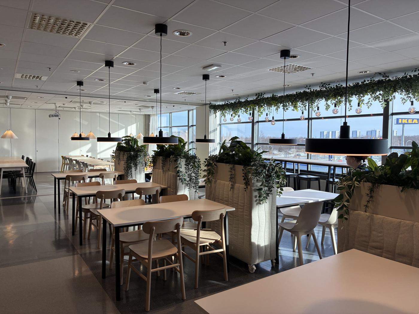 Ljus och modern personalmatsal med träbord, hänglampor, gröna växter och stora fönster med utsikt över en IKEA-skylt.