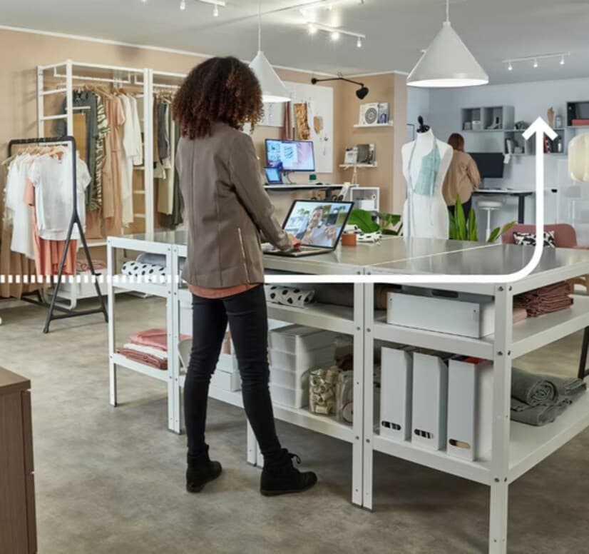 Ljust arbetsrum med klädställ, person vid laptop, stora lampor i taket och modeartiklar i bakgrunden.