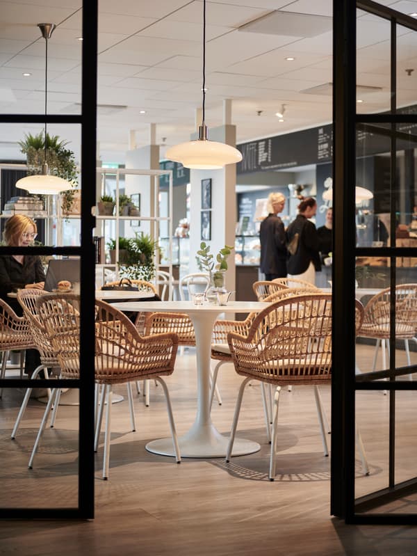 Ljust caféinteriör sett genom glasdörrar, med runda bord, rottingstolar, hängande lampor och en disk i bakgrunden där kunder står.