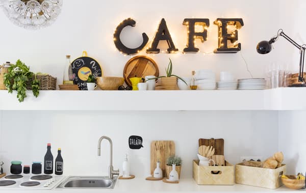 Lletres il·luminades que escriuen «cafe» a la paret sobre un prestatge llarg i el taullel i l'aigüera d'aquesta cuina blanca.
