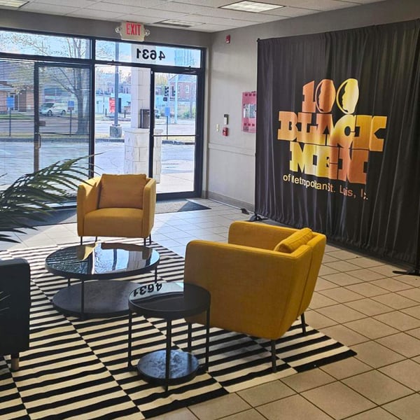 Lobby with yellow chairs, a striped rug, and a backdrop reading ‘100 Black Men’ near large front windows.
