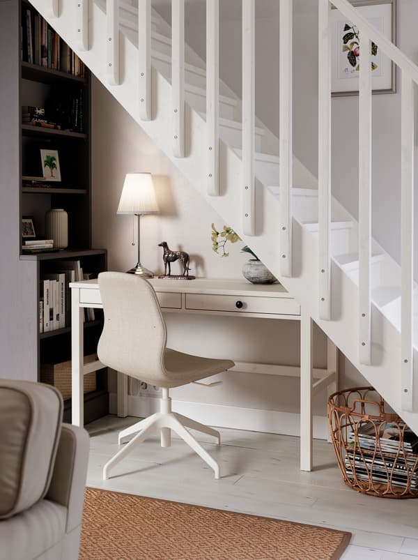 Located under a staircase is a workspace consisting of a white HEMNES desk, a LÅNGFJÄLL chair, a lamp and a tall bookcase.