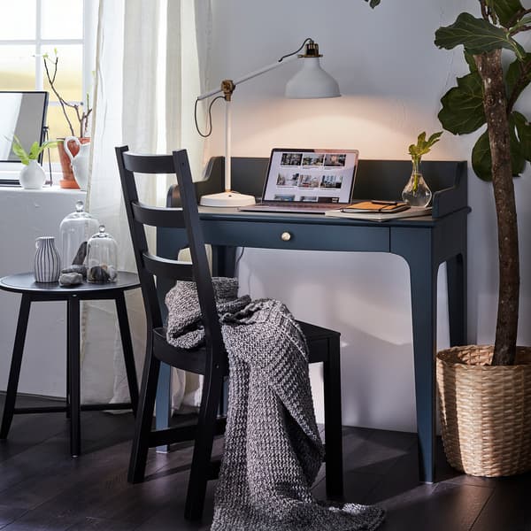 LOMMARP dark blue-green desk has a laptop and a lit table lamp on it and in front of it is a black chair with a gray throw.