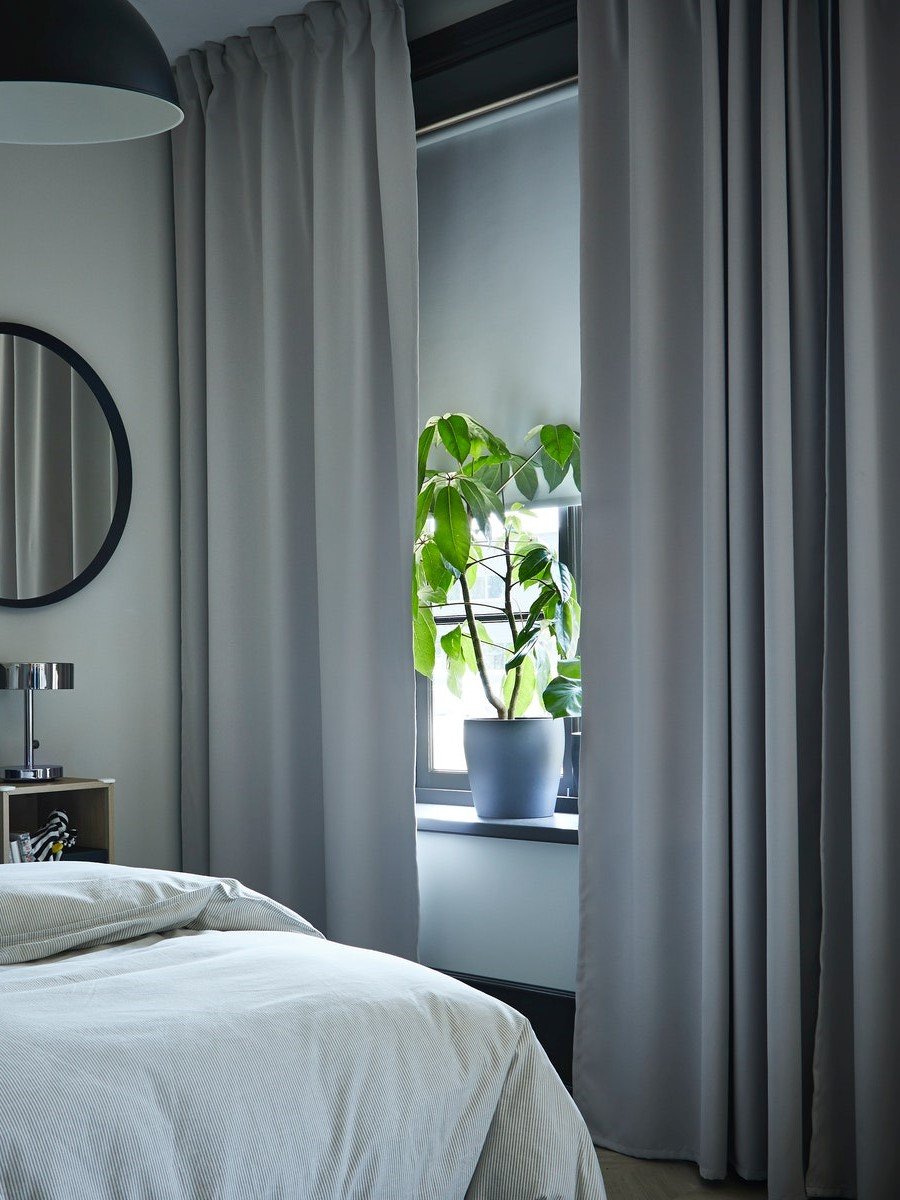 Ložnice s šedými zatemňovacími závěsy IKEA, černým kulatým zrcadlem na stěně, stolkem s lampou a zelenou pokojovou rostlinou v květináči na parapetu.