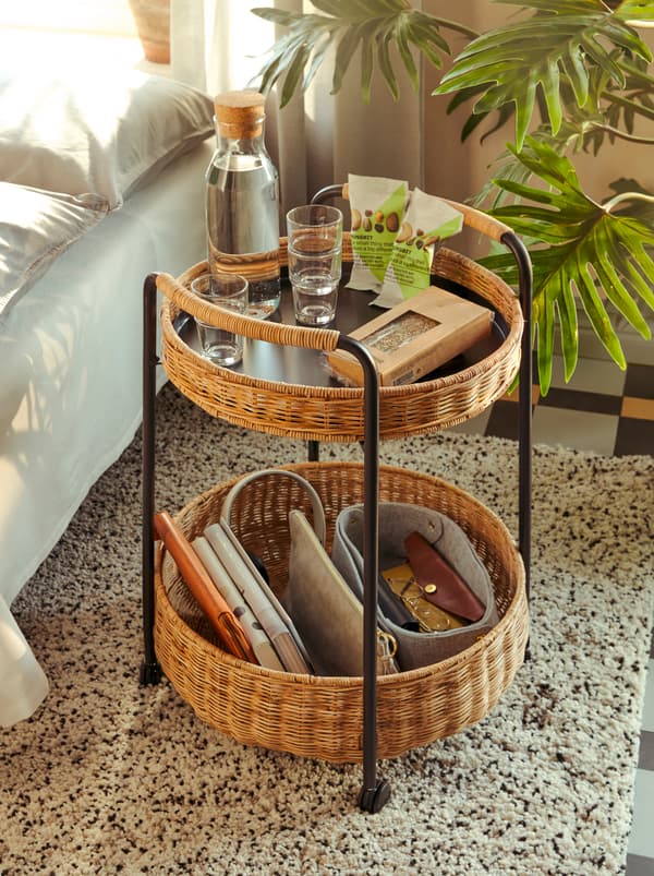 รถเข็นทำจากหวาย LUBBAN/ลูบบัน ชั้นหนึ่งใส่ขนมขบเคี้ยว และขวดแก้ว IKEA 365+/อิเกีย 365+ มีน้ำอยู่ รวมทั้งของใช้ส่วนตัวอื่น ๆ