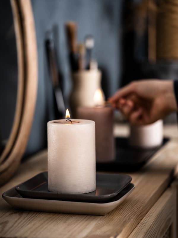 ตู้ไซด์บอร์ดทำจากไม้ในห้องนอนที่มีกระจกเงาและของตกแต่ง มือข้างหนึ่งจุดเทียนหอม LUGGA/ลุกก้า สองเล่มที่วางอยู่บนตู้ด้วย