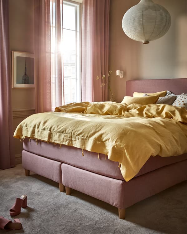 Lumière matinale dans une chambre à coucher avec un lit-sommier brun clair-rose, une housse de couette jaune et une suspension blanche ronde.